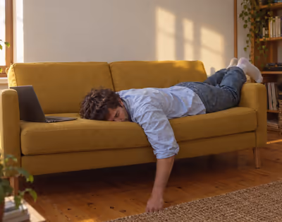 A man lying face down on a mustard sofa, his laptop open on the armrest